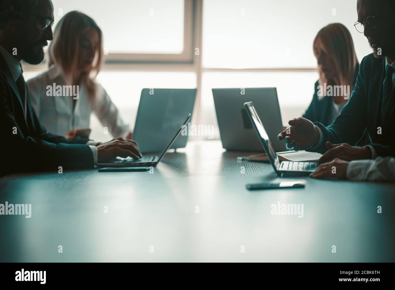Silhouetten von Geschäftsleuten, die am Computer arbeiten, während sie symmetrisch am Bürotisch vor dem Hintergrund eines sonnendurchfluteten Fensters sitzen. Unternehmen Stockfoto
