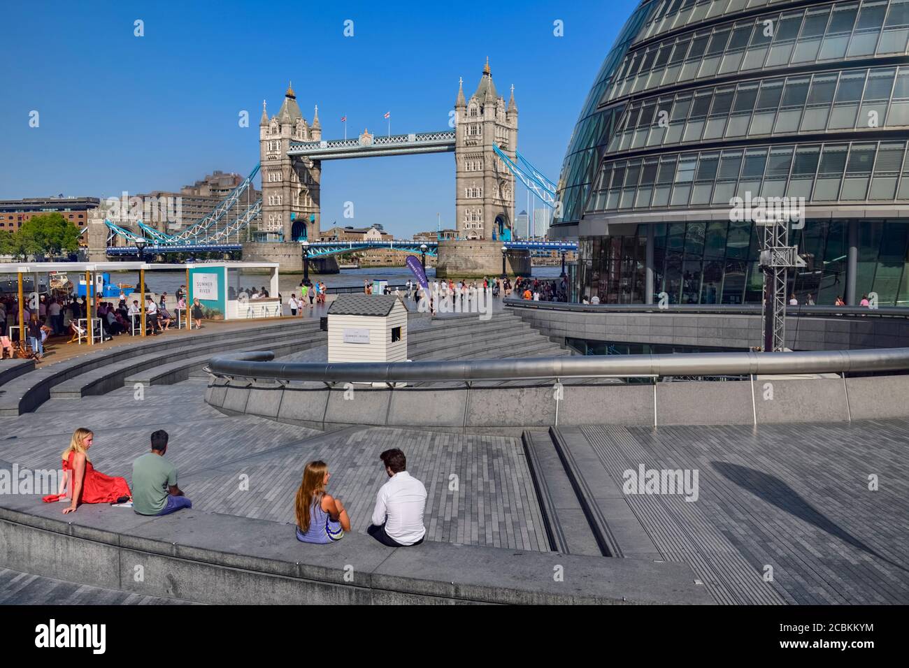 England, London, Tower Bridge mit Leuten, die die Sommersonne im Scoop genießen. Das ist ein versunkenes Amphitheater neben dem Rathaus. Stockfoto