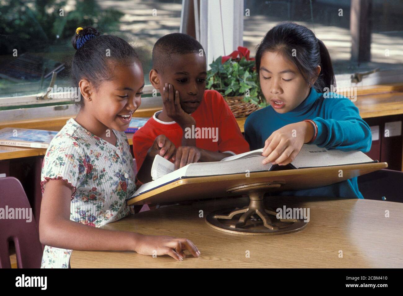 Austin, Texas, USA. 1990: Ethnisch gemischtes Trio von Grundschülern, die in der Schulbibliothek Wörter in einem großen, ungekürzten Wörterbuch nachschlagen. Modellversion. ©Bob Daemmrich Stockfoto