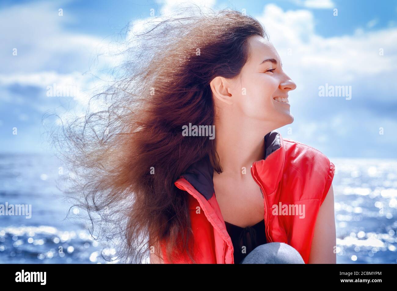 Lächelnde Frau in einer roten ärmellosen Jacke sitzt am Meer mit langen lockigen Haaren fliegen im Wind. Outdors Profil Hochformat Stockfoto