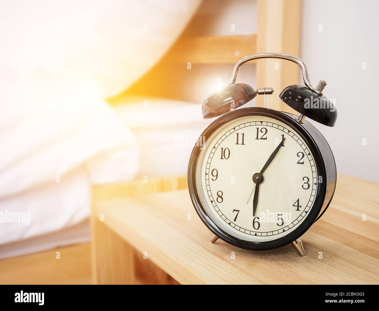 Wecker, Weckzeitkonzept: Retro-Wecker mit fünf Minuten nach sechs Uhr morgens auf Holzbett Beistelltisch mit weißem Bettlaken Stockfoto