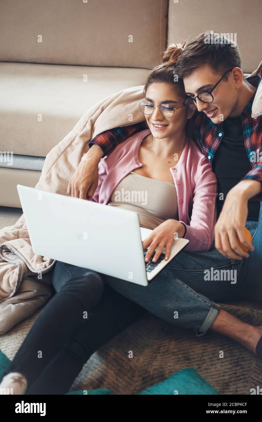 Junge kaukasische Paar zusammen auf dem Boden in der Nähe der liegen Sofa mit einem Laptop mit Deckchen tragen Brillen und bedeckt Lächeln Stockfoto