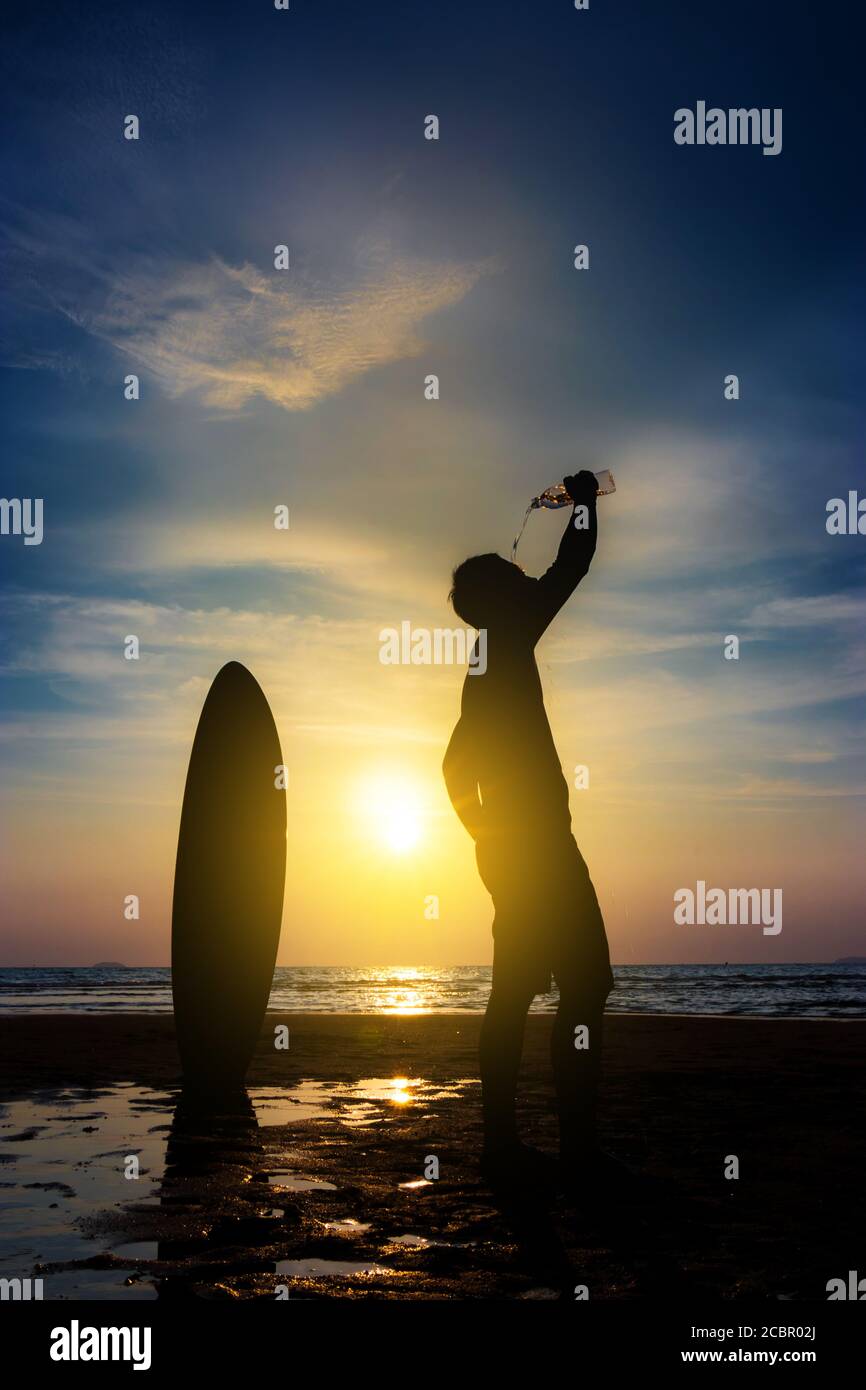 Silhouette von Surf Mann stehen mit einem Surfbrett Wasser aus Flasche trinken. Surfen am Strand bei Sonnenuntergang. Outdoor Wassersport Abenteuer Lifestyle. Sommersport Stockfoto