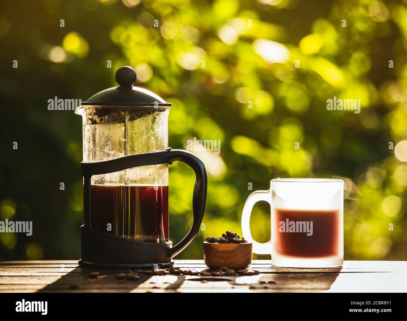 Französische Presse Kaffee und Tasse auf dem Tisch mit verstreuten Kaffeebohnen im Garten an sonnigen hellen Morgen Tag. Grund zum Aufwecken Konzept. Bokeh grün. Stockfoto