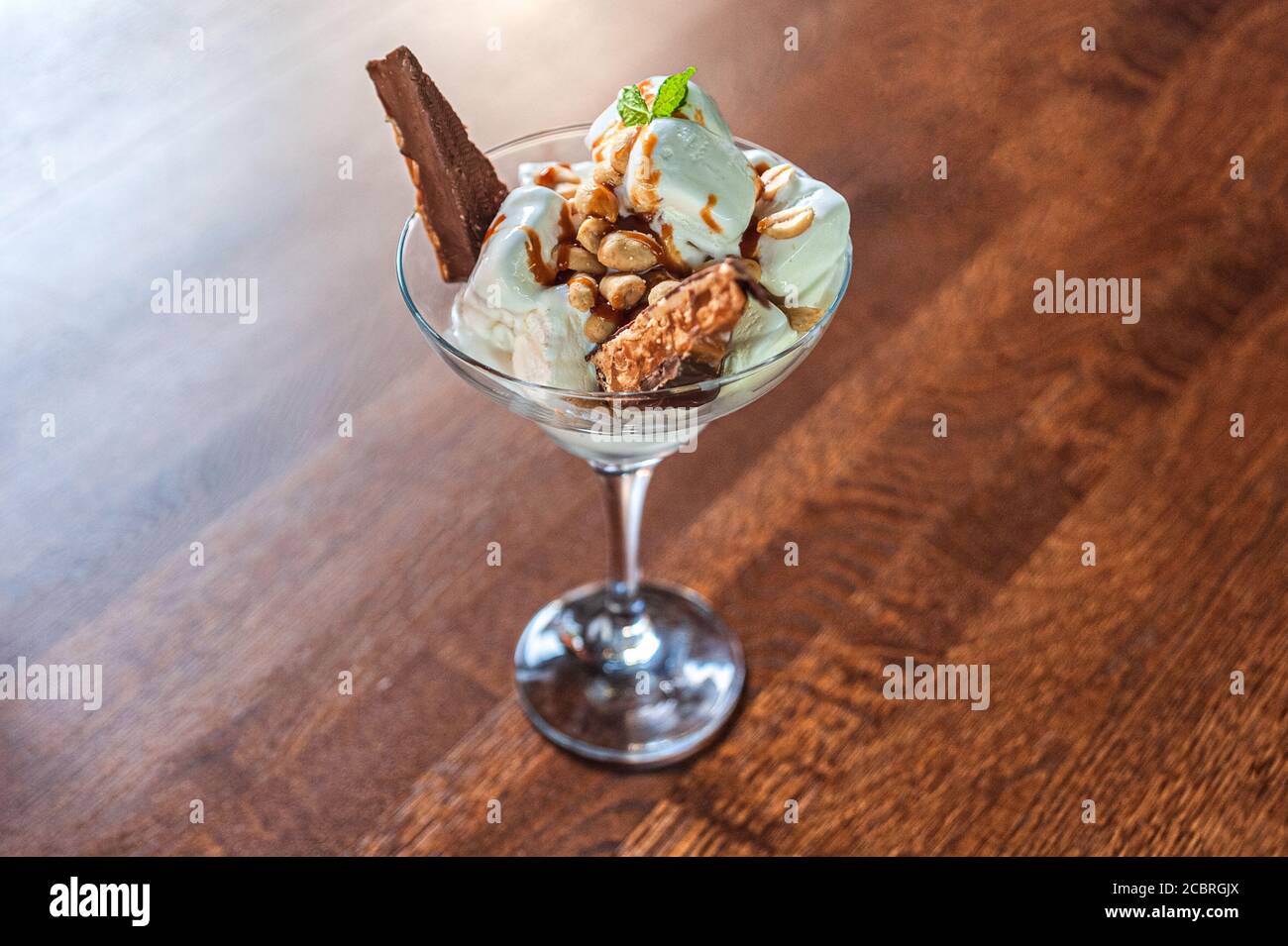 Dessert aus weißem Eis, Erdnüssen und einer Schokoladenriegel mit Minzblatt in einer Glasschale auf einem Holztisch Stockfoto