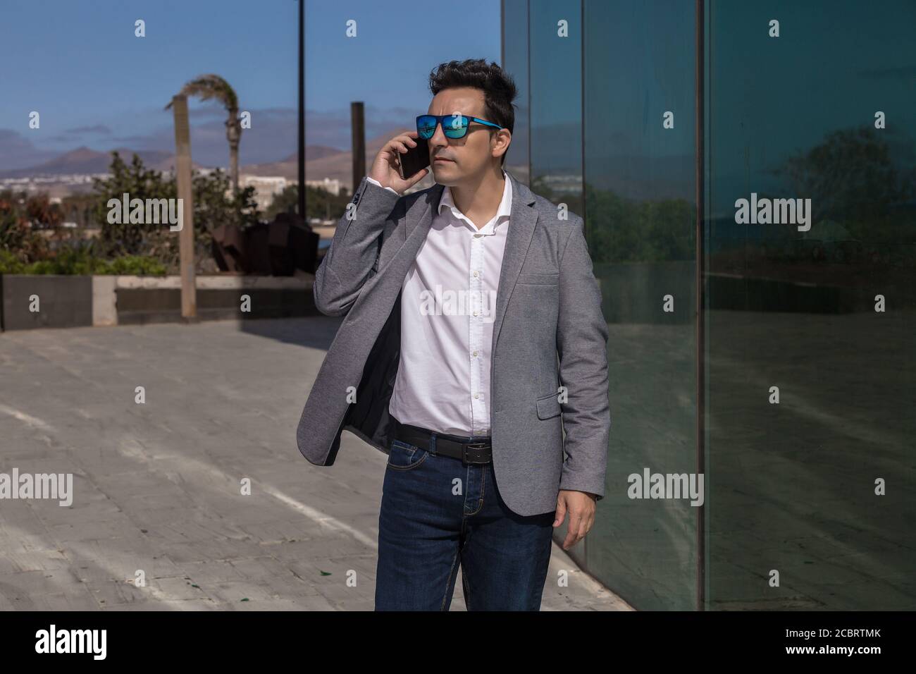 Erwachsene Mann in eleganten Freizeitkleidung und Sonnenbrille wegschauen Und mit Smartphone-Gespräch in der Nähe der Glaswand auf der Straße der Stadt Stockfoto