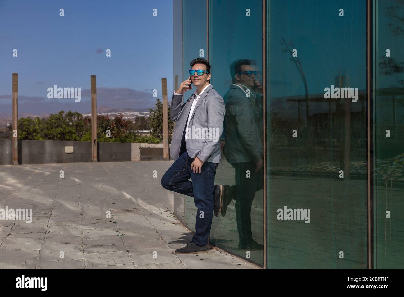 Ganzkörper froh männlichen Unternehmer in smart casual Outfit lächelnd Und mit Smartphone-Gespräch, während lehnte sich an Glas Gebäudewand An der City Street Stockfoto