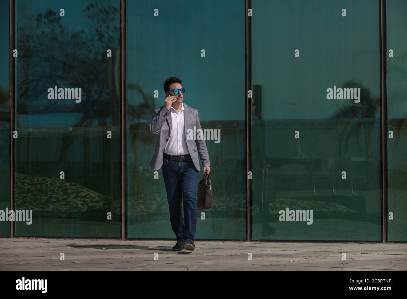 Full Body serious Erwachsenen Mann in smart casual Outfit suchen Unterwegs und auf dem Smartphone sprechen, während Sie in der Nähe der Glaswand gehen An der City Street Stockfoto