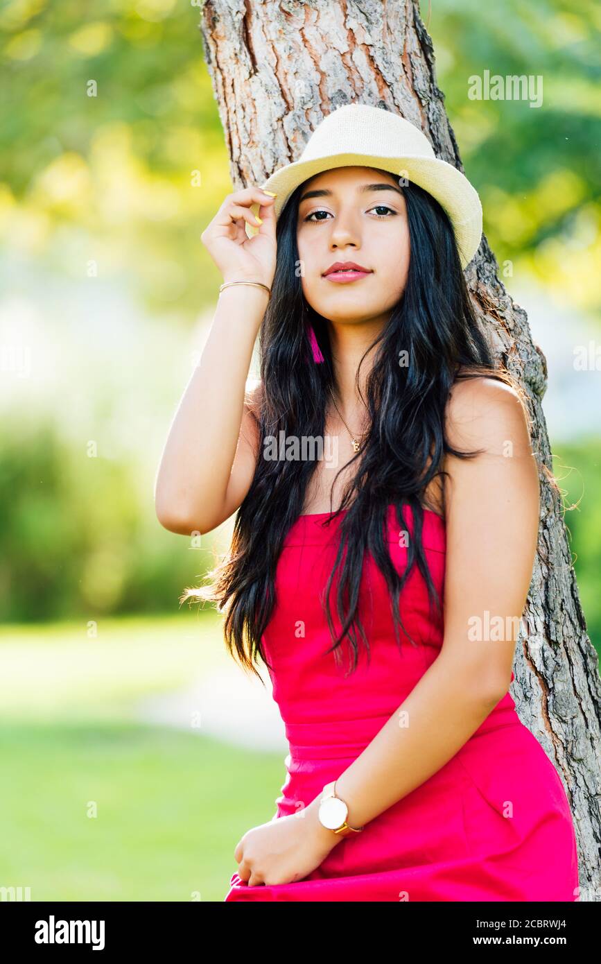 Junge Brünette Latina Mädchen mit langen schwarzen Haaren und schwarzen Augen, trägt ein rotes Kleid und leichten Hut, Blick auf die Kamera auf einem Baumstamm lehnt. Stockfoto