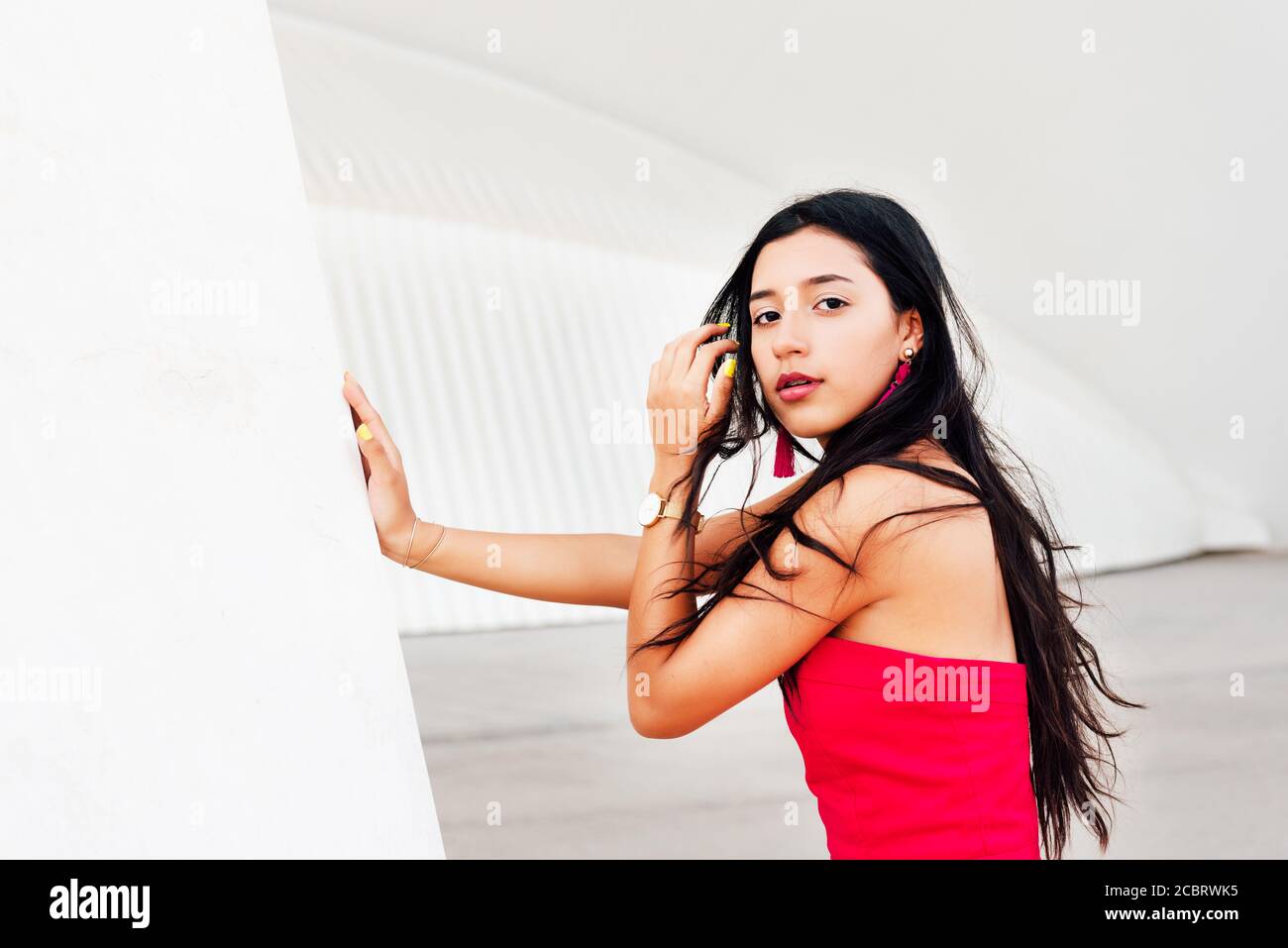 Portrait von eleganten lateinischen jungen Mädchen mit langen Brünette Haare Und rotes Kleid lehnte mit einer Hand auf weißem Rahmen Und die Kamera mit unordentlichen Haaren betrachten Stockfoto