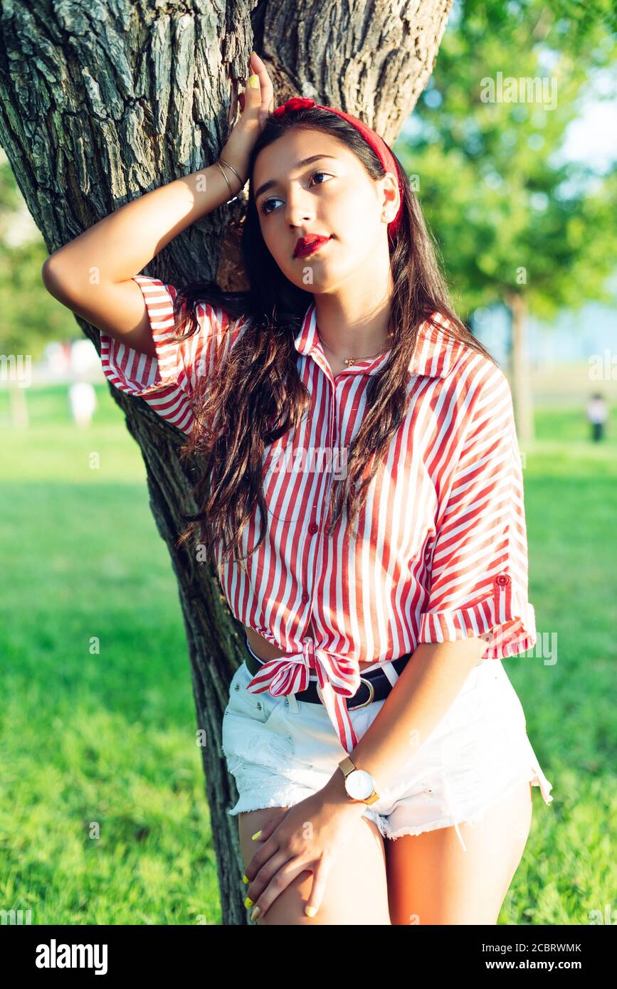 Junge lateinische Mädchen, langhaarige Brünette, trägt ein Hemd aus weißen und braunen Streifen und roten Band in den Haaren, lehnte sich auf einen Baumstamm Stockfoto