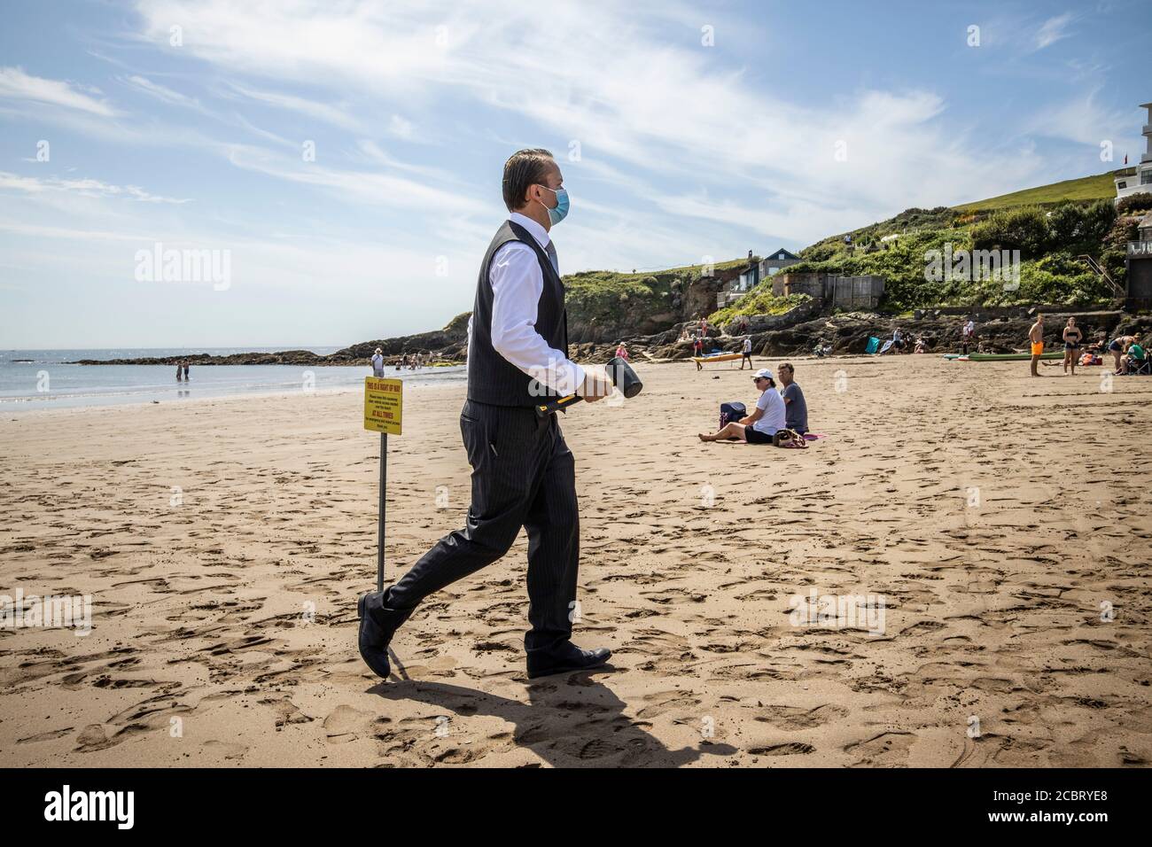 Ein Hotelangestellter vom Burgh Island Hotel geht über die Strand trägt eine schützende Gesichtsmaske und Anzug, wie er Markiert die Servicestrecke über die Bea Stockfoto