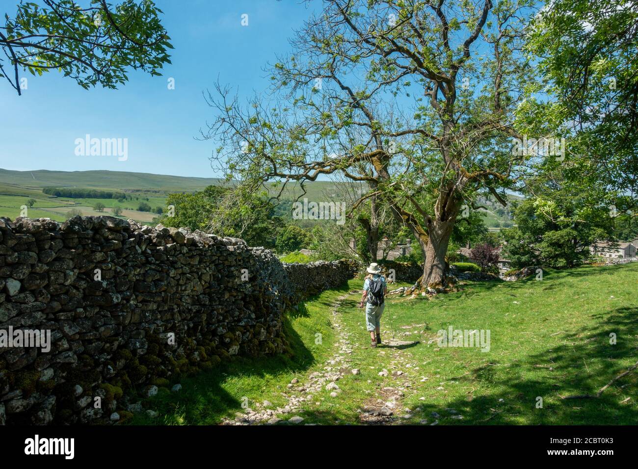 Landschaft in Großbritannien: Weibliche Wanderer, die an einem heißen Sommertag, Yorkshi, durch ein grünes Grasfeld an einer alten Steinscheune auf dem Grassington Woods Walk vorbeigehen Stockfoto