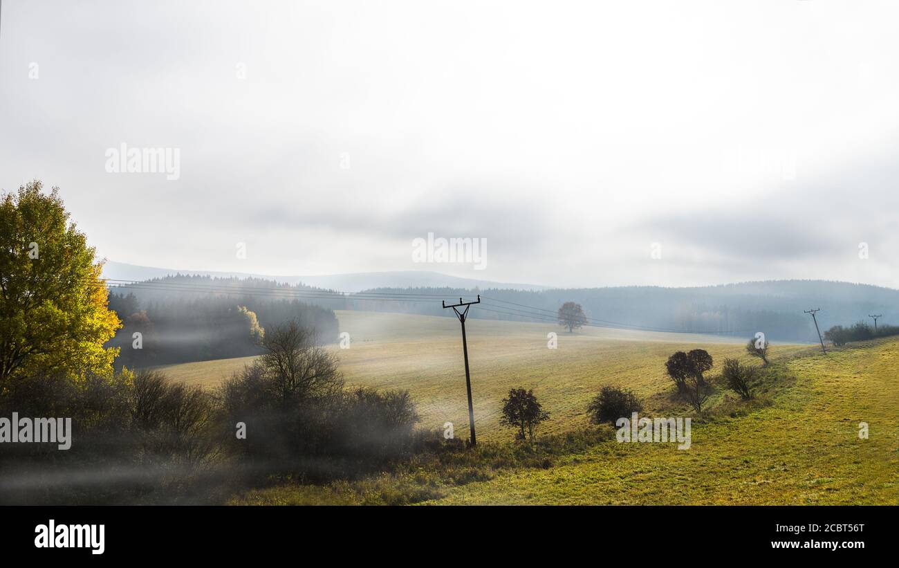 Landschaftlich reizvolle Herbstlandschaft mit Stromleitung und Nebel über Wiesen und bewaldeten Hügeln. Melancholisches Grünlandpanorama mit alten Strommasten. Stockfoto