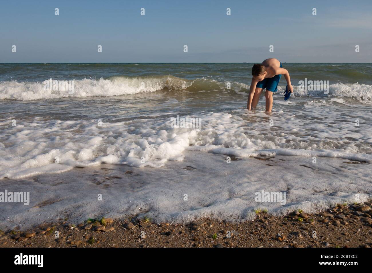Ein 10-jähriger Junge zieht an einem sonnigen Tag Hausschuhe am Meer an. Stockfoto