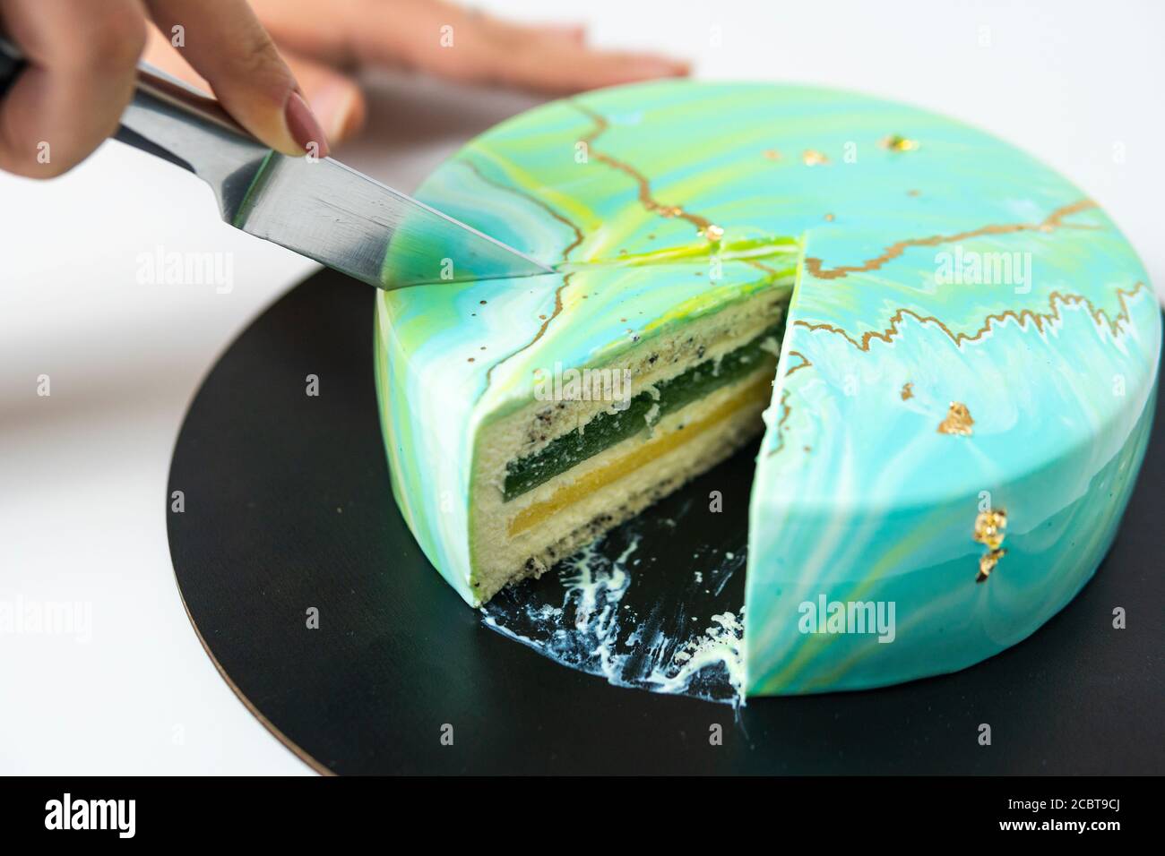Nahaufnahme des Schneidens mit Messer Mousse Kuchen bedeckt blau und grün Spiegelglasur. Französisches Dessert. Gefrorener Spiegel Sahnehäubchen auf dem Kuchen. Backen und Konfektion Stockfoto