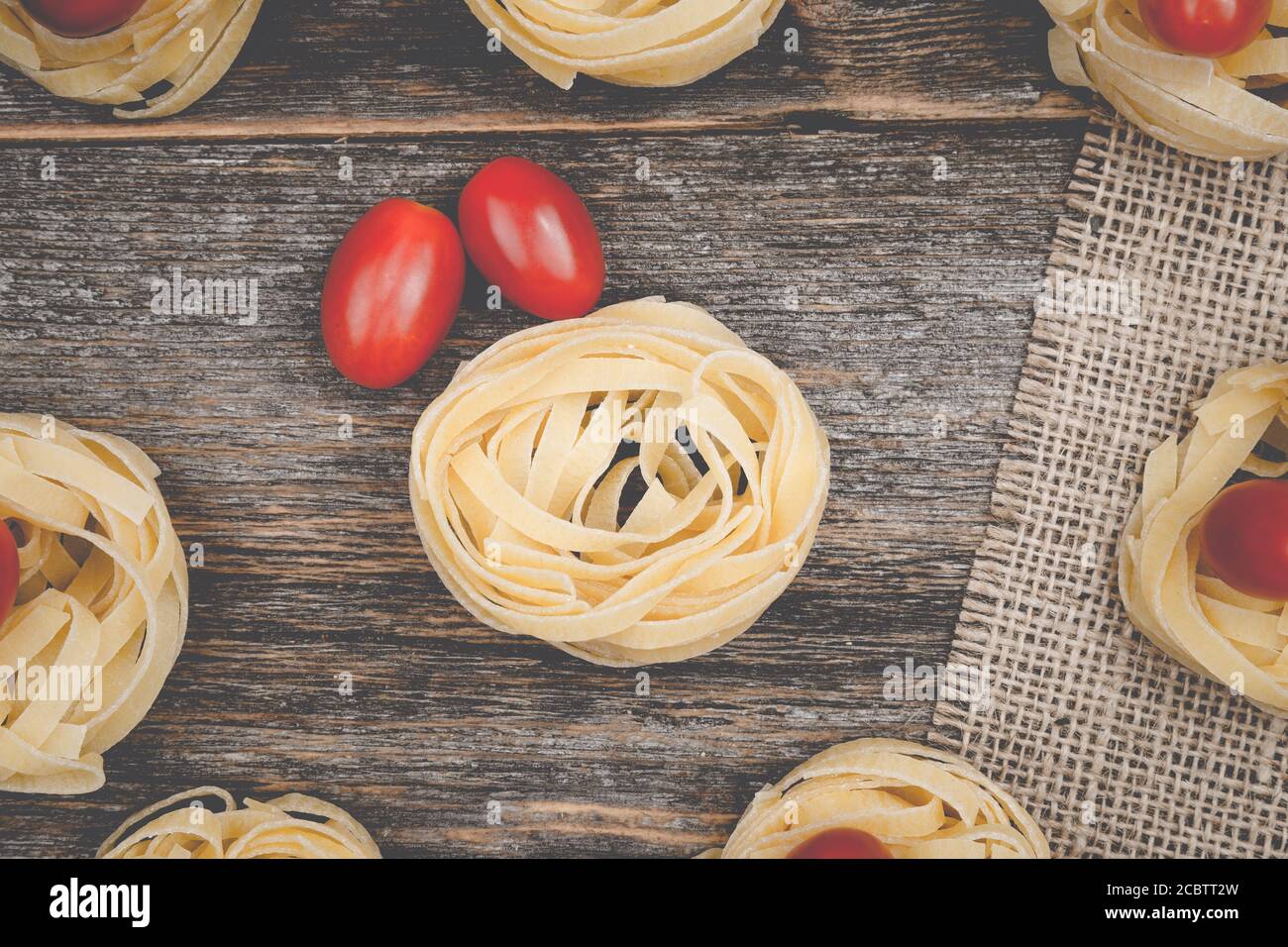 Nahaufnahme von Tagliatellennestern mit eiförmigen Mini-roma-Tomaten auf einem rustikalen Holztisch mit Sackleinen, Stockfoto