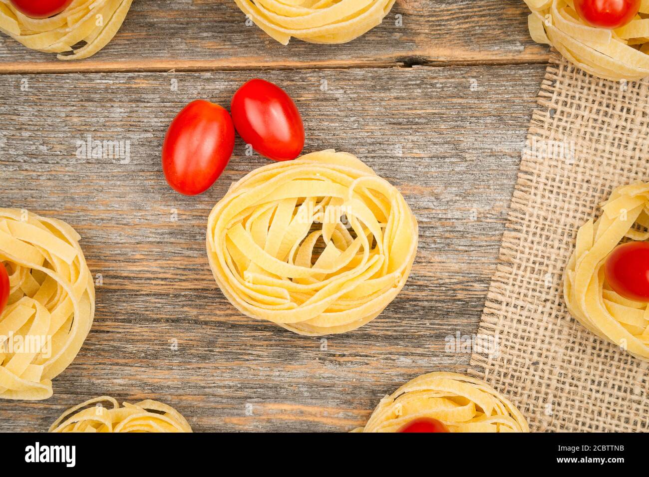Nahaufnahme von Nestern von Tagliatelle Nudeln mit Ei-förmigen Mini roma Tomaten auf einem rustikalen Holztisch mit Sackleinen, Stockfoto