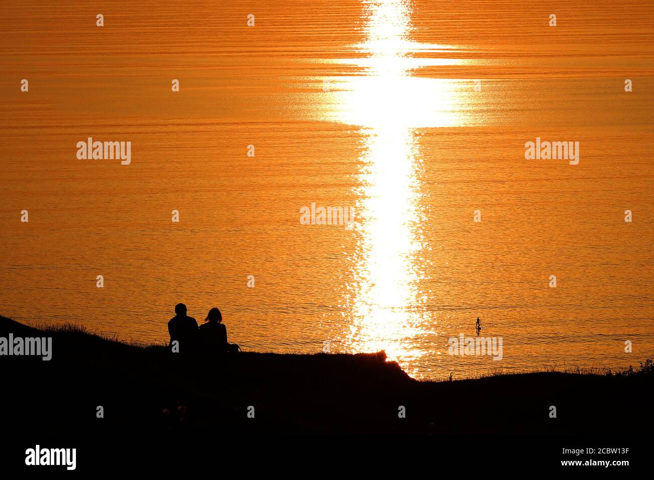 Ein Paddelboarder fährt durch das ruhige Wasser, während ein Paar den Sonnenuntergang an der Rhossili Bay im Gower South Wales an einem wunderschönen Sonnenuntergang-Abend beobachten kann Stockfoto