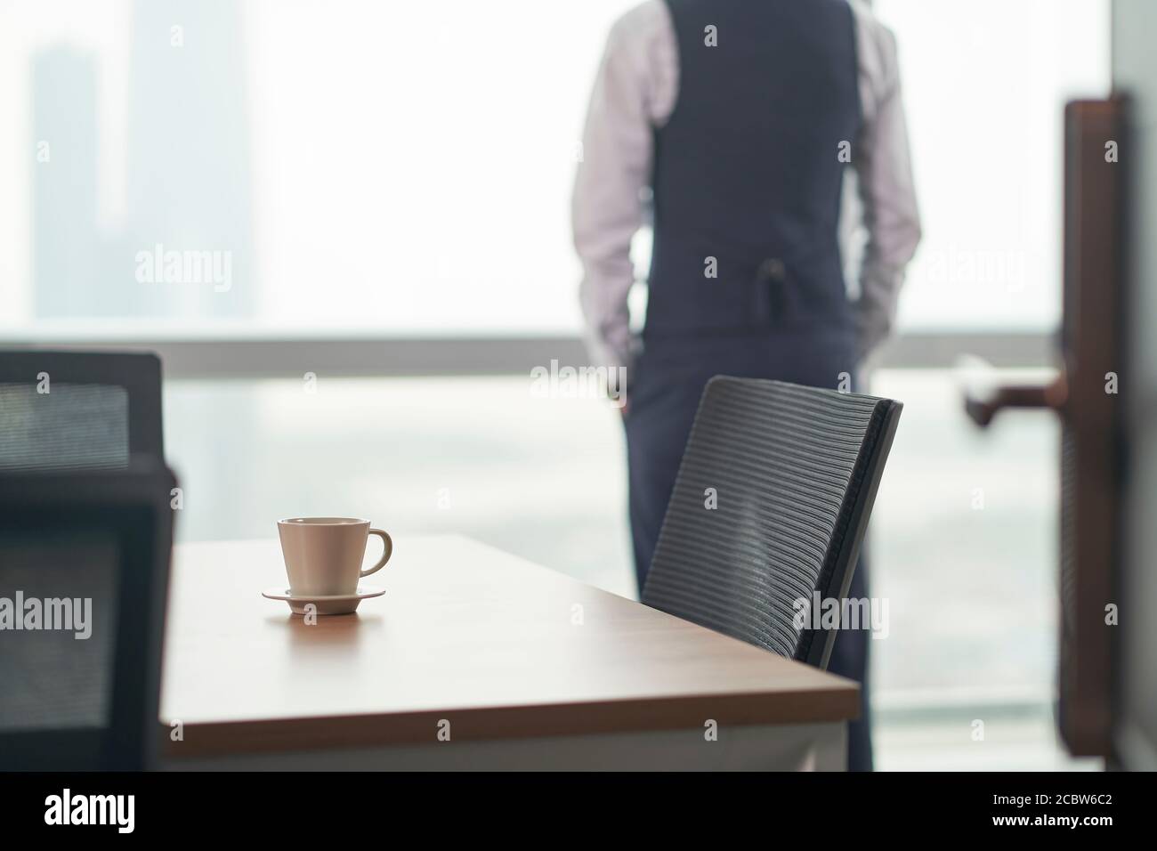 asiatischer Geschäftsmann steht vor dem Fenster im Büro denken, Hände in den Taschen, Fokus auf Vordergrund Stockfoto