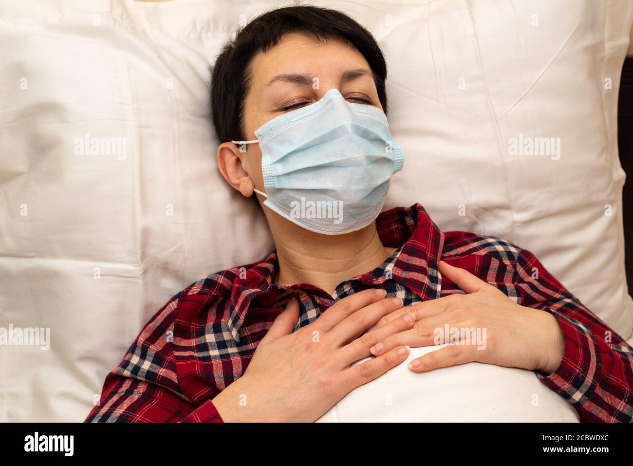 Kranke junge Frau in medizinischer Maske im Bett liegend Gefühl Brustschmerzen und keuchend. Kurzhaarige Brünette mit Symptomen von covid-19 oder Lungenentzündung zu Hause Stockfoto