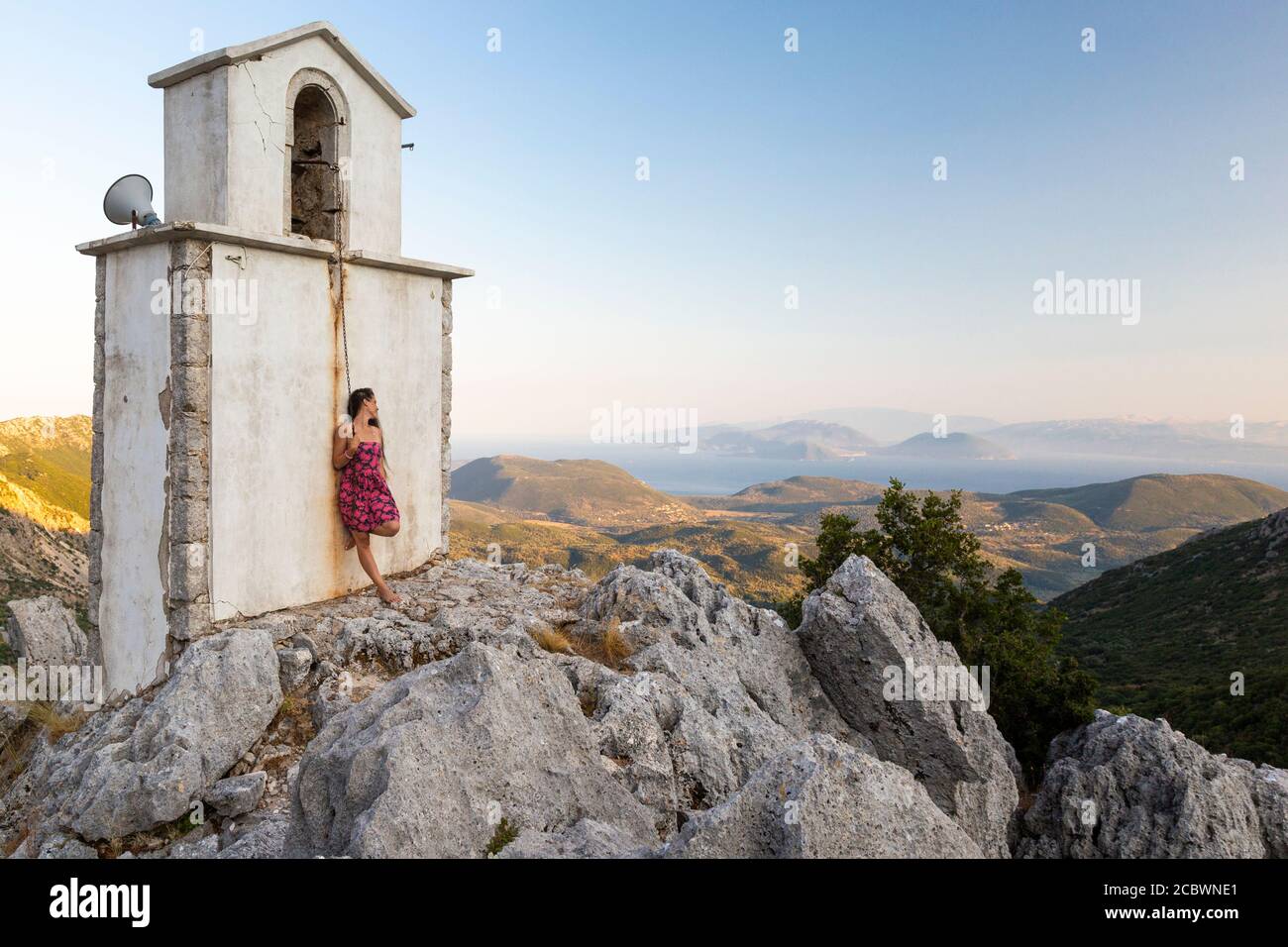 Ein Mädchen lehnt sich an einen Glockenturm in den Bergen und genießt die Aussicht in der Nähe von Cerkev Agia Paraskevi, Lefkada, Ionische Inseln, Griechenland Stockfoto