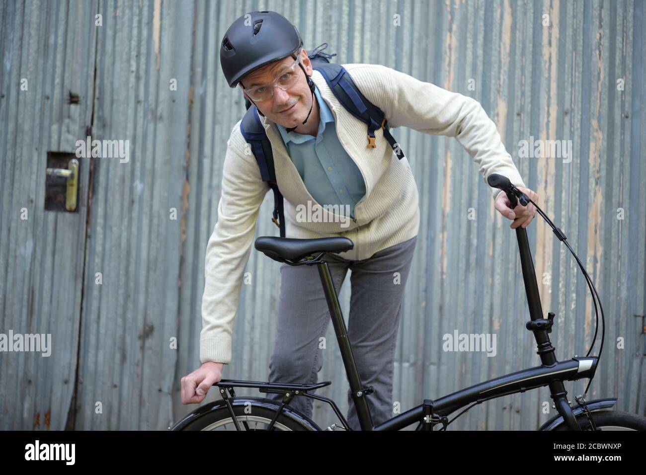 Reifer Kaukasischer Mann in einem Fahrradhelm mit seinem Fahrrad Gegen eine graue Tür Stockfoto