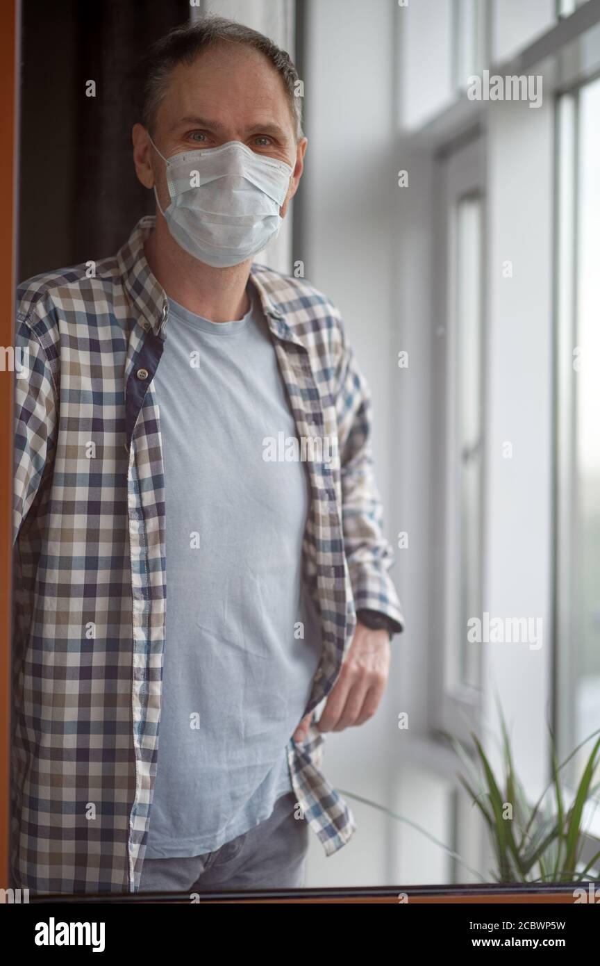 Reife kaukasischen Mann in medizinischen Gesichtsmaske Aufenthalt an der Fenster seiner Wohnung Stockfoto