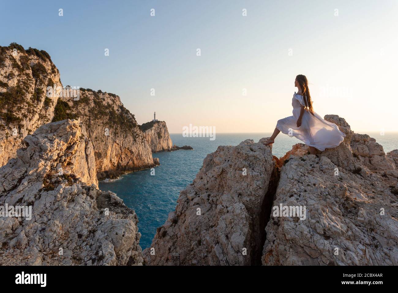 Ein Mädchen in weißem Kleid blickt auf den Leuchtturm an der Spitze des Kap Lefkada bei Sonnenuntergang, Lefkada, Ionische Inseln, Griechenland Stockfoto