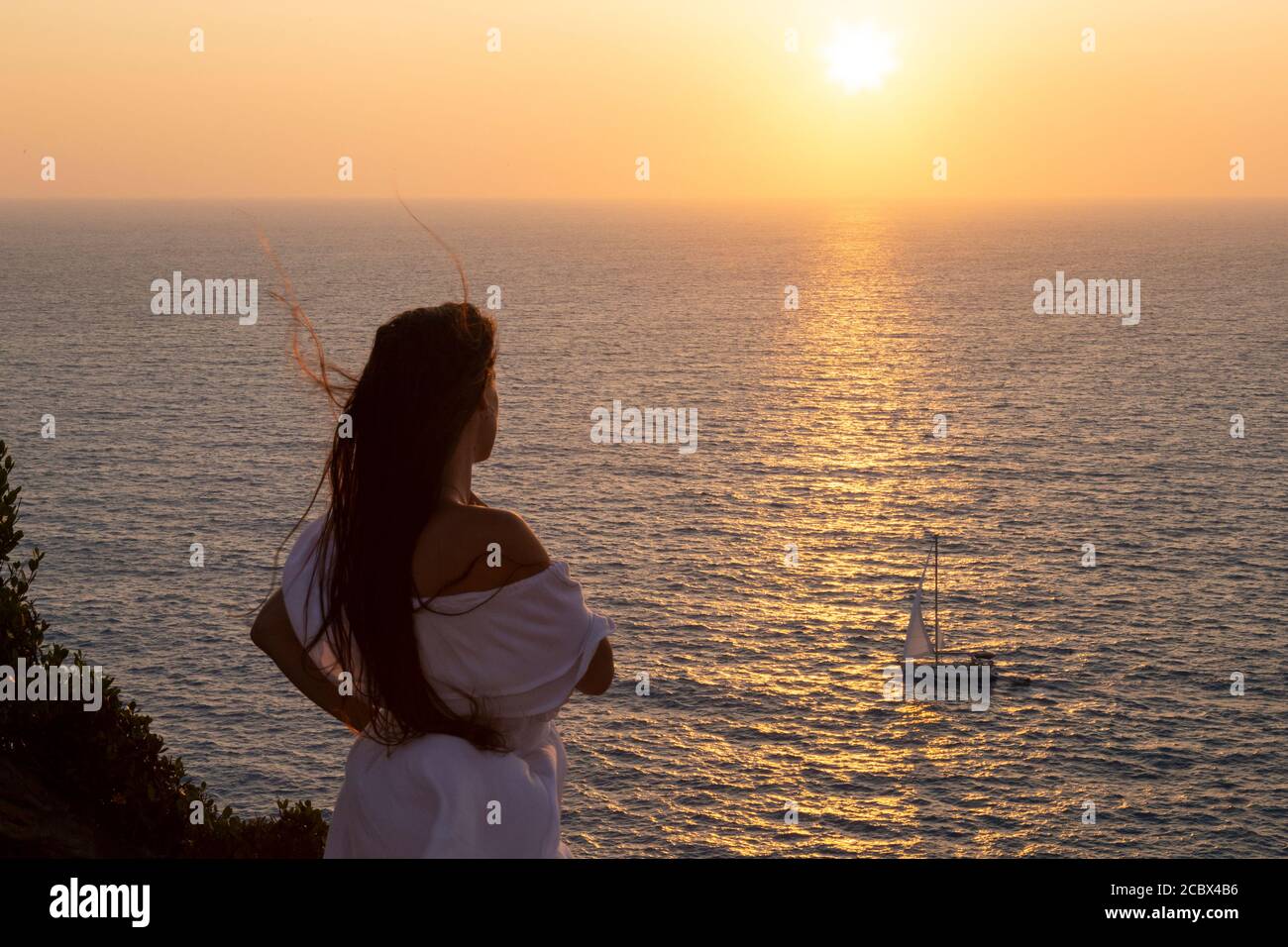 Ein Mädchen in weißem Kleid sieht einem Segelboot vorbei bei Sonnenuntergang, Kap Lefkada, Lefkada, Ionische Inseln, Griechenland Stockfoto