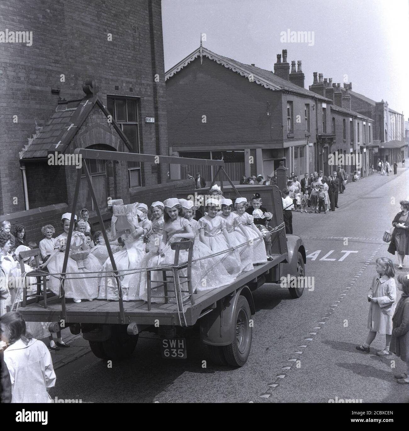 1950er Jahre, historisch, Rose Queen Feiern....Einheimische beobachten einen offenen LKW, der entlang der Market Street in Farnworth, Bolton, Lancashire, England, Großbritannien fährt, Sie tragen junge Mädchen in ihren Kleidern und nehmen an dem jährlichen Rose Queen Festival Teil. Normalerweise findet es im Juni statt und geht auf die 1880er Jahre zurück. Nach den Kriegen der Rosen (1455–87) wurde es zu einem bedeutenden jährlichen Ereignis in Städten und Dörfern in ganz Großbritannien, insbesondere in Lancashire, dem sogenannten Red Rose County. Stockfoto