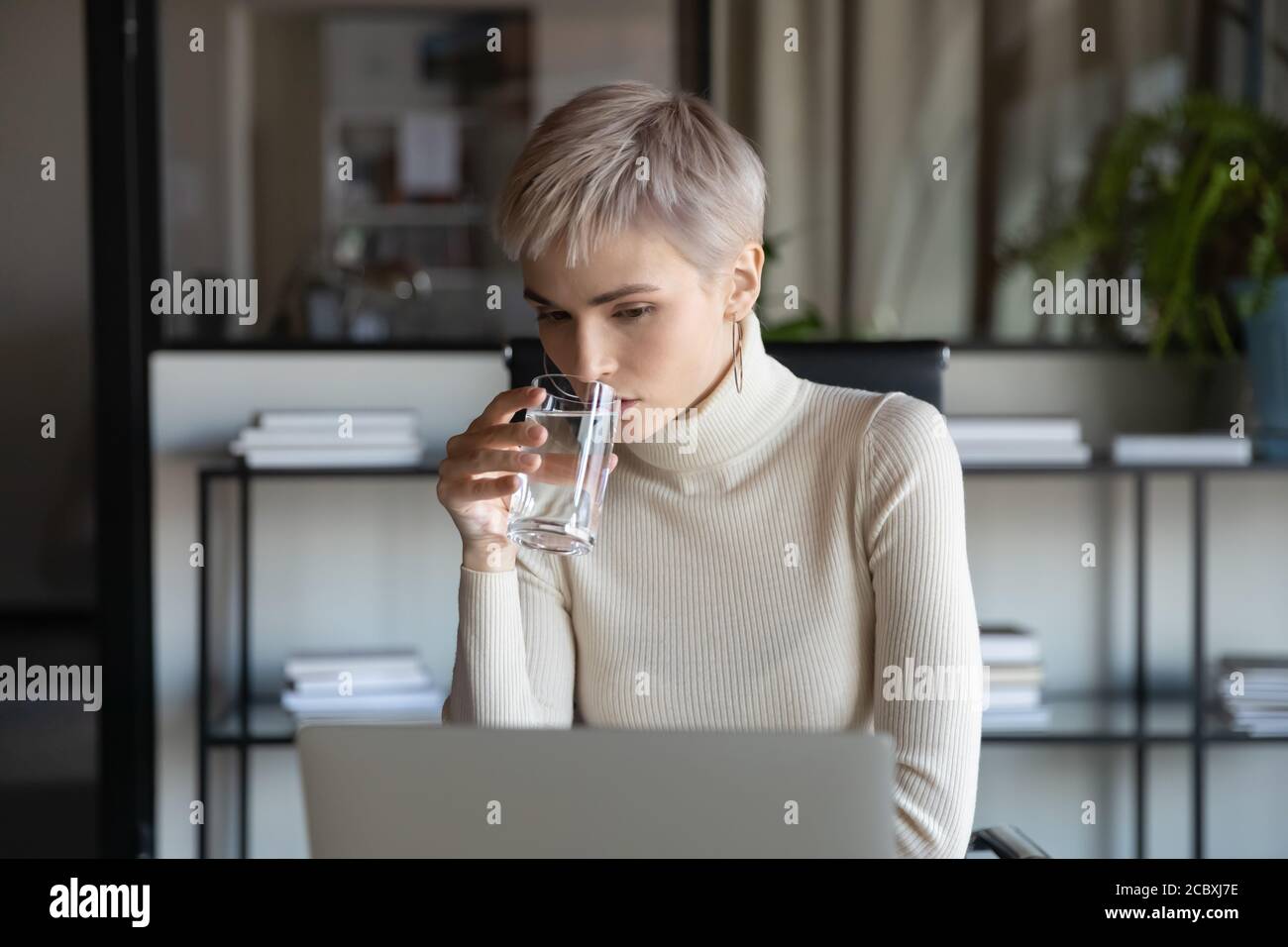 Fokussierte junge blonde Geschäftsfrau Trinkwasser am Arbeitsplatz. Stockfoto