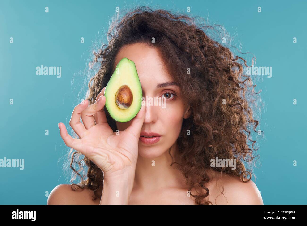 Junge Frau, die Sie mit dem linken Auge anschaut, während sie hält Avocado von einem anderen Stockfoto
