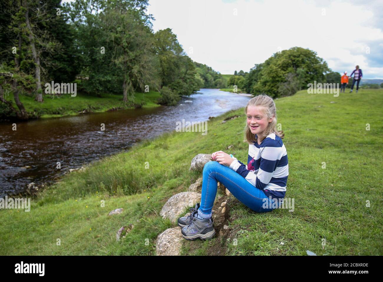 Mädchen sitzt in der Nähe von River Wharfe, Grassington, während die Familie in der Nähe spazieren geht Stockfoto