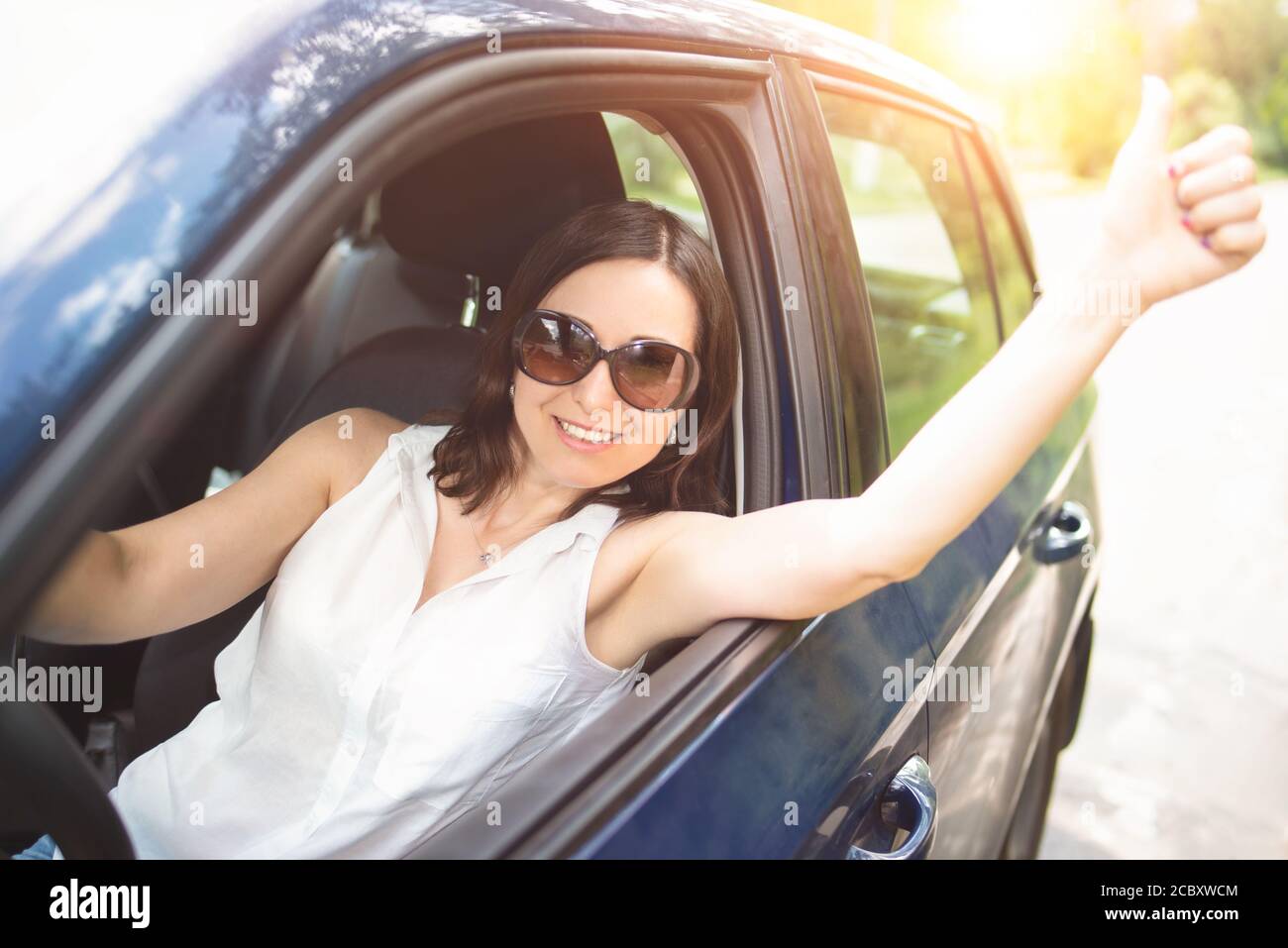 Eine elegant aussehende Frau mittleren Alters mit Sonnenbrille legte ihre Hand aus dem Autofenster und hob ihre Daumen nach oben. Stockfoto