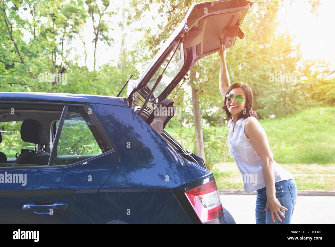 Lächelnde kaukasische Frau in einer weißen Bluse und Denim-Shorts Ihre Stofftaschen in den Kofferraum des Autos stecken Stockfoto
