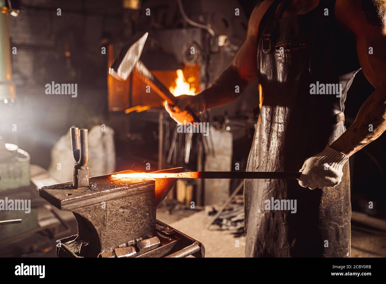 Muskulöser Erwachsener mit Bart trägt Lederschürze in der Werkstatt, Heizung. Gehärtetes Metall im Ofen Stockfoto