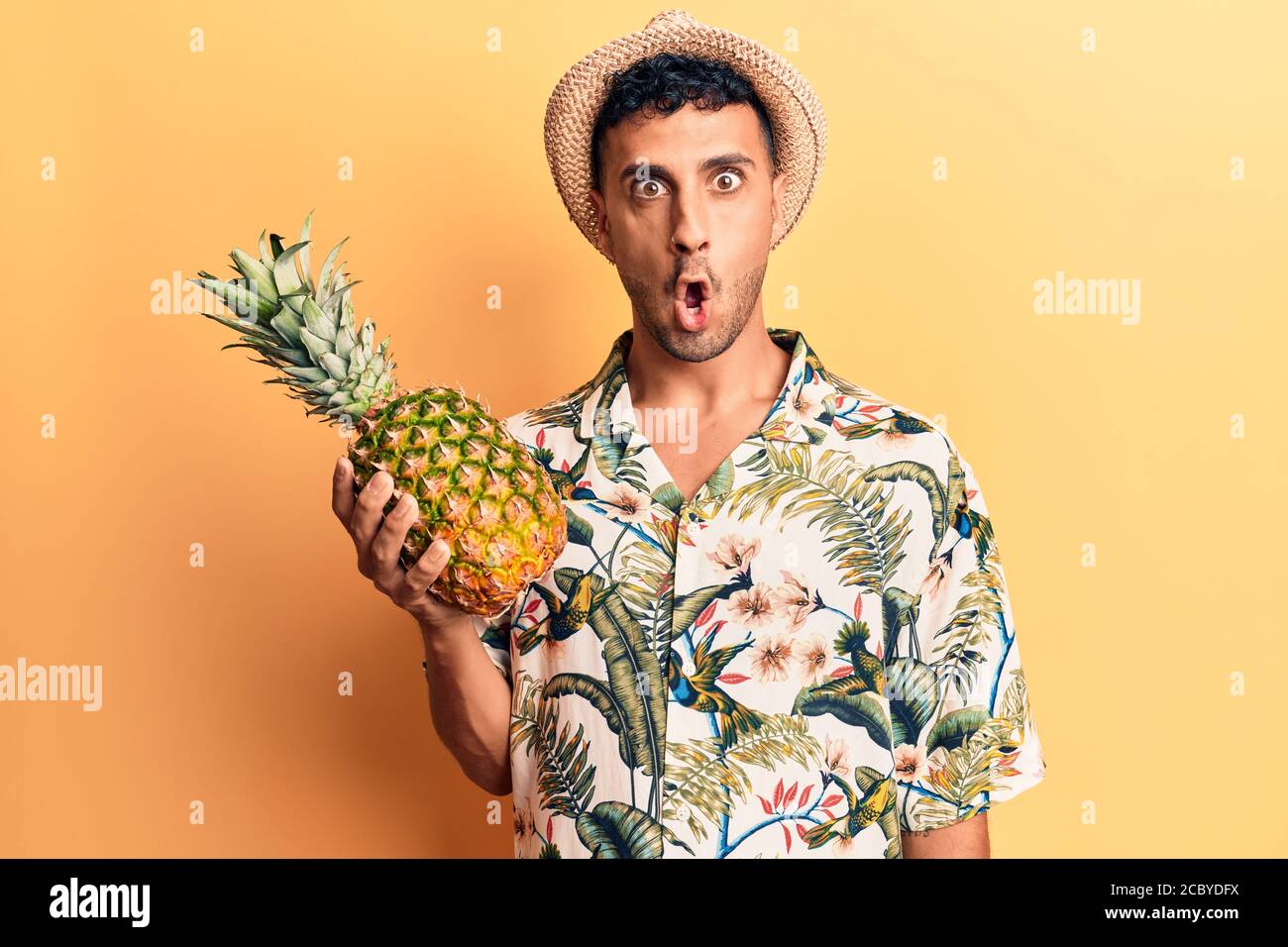 Junger hispanischer Mann trägt Sommerhut mit Ananas verängstigt und erstaunt mit offenem Mund