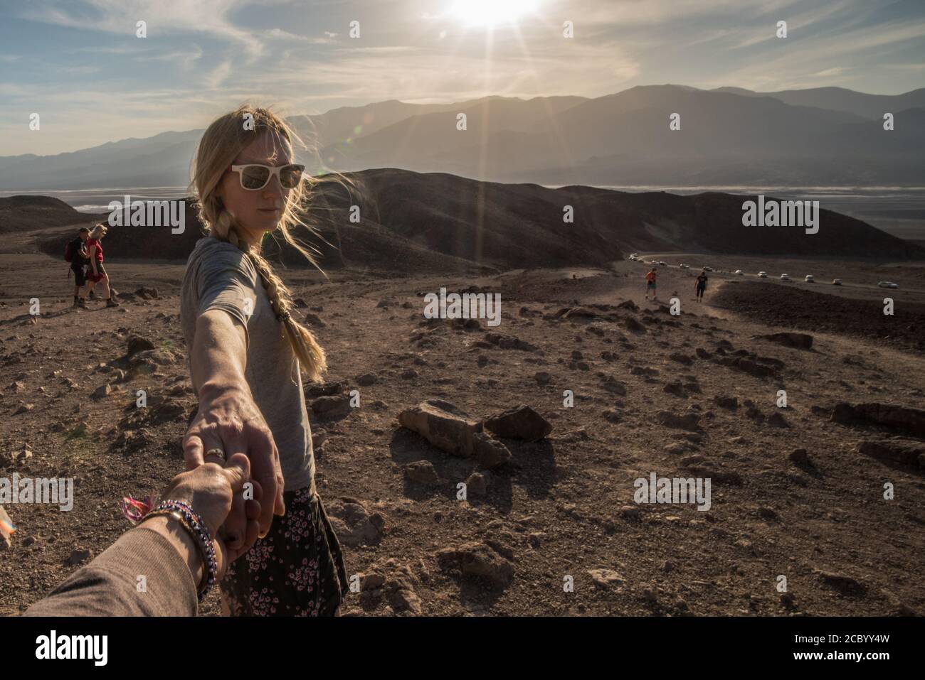 Die blonde Wanderin hält die Hände der Fotografin und blickt zurück in die Kamera, während ihr Haar weht und die Sonne im Death Valley Nationalpark untergeht. Stockfoto