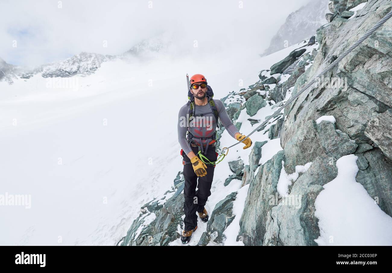 Männlicher Alpinist in Sonnenbrille und Schutzhelm, der festsitzend Seil hält, während er verschneiten Berg klettert. Bergsteiger steigt natürliche Felsformation. Konzept des Winterkletterns. Stockfoto
