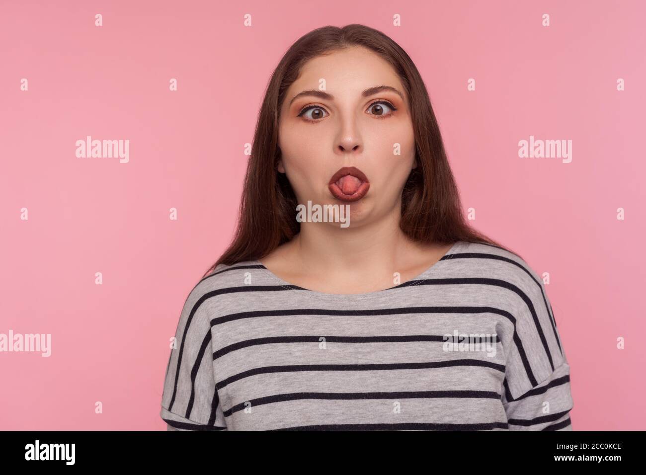 Portrait einer lustigen dummen amüsanten Frau im gestreiften Sweatshirt, die mit gekreuzten Augen steht, die Zunge zeigt und einen dummen, hirnlosen Gesichtsausdruck macht Stockfoto
