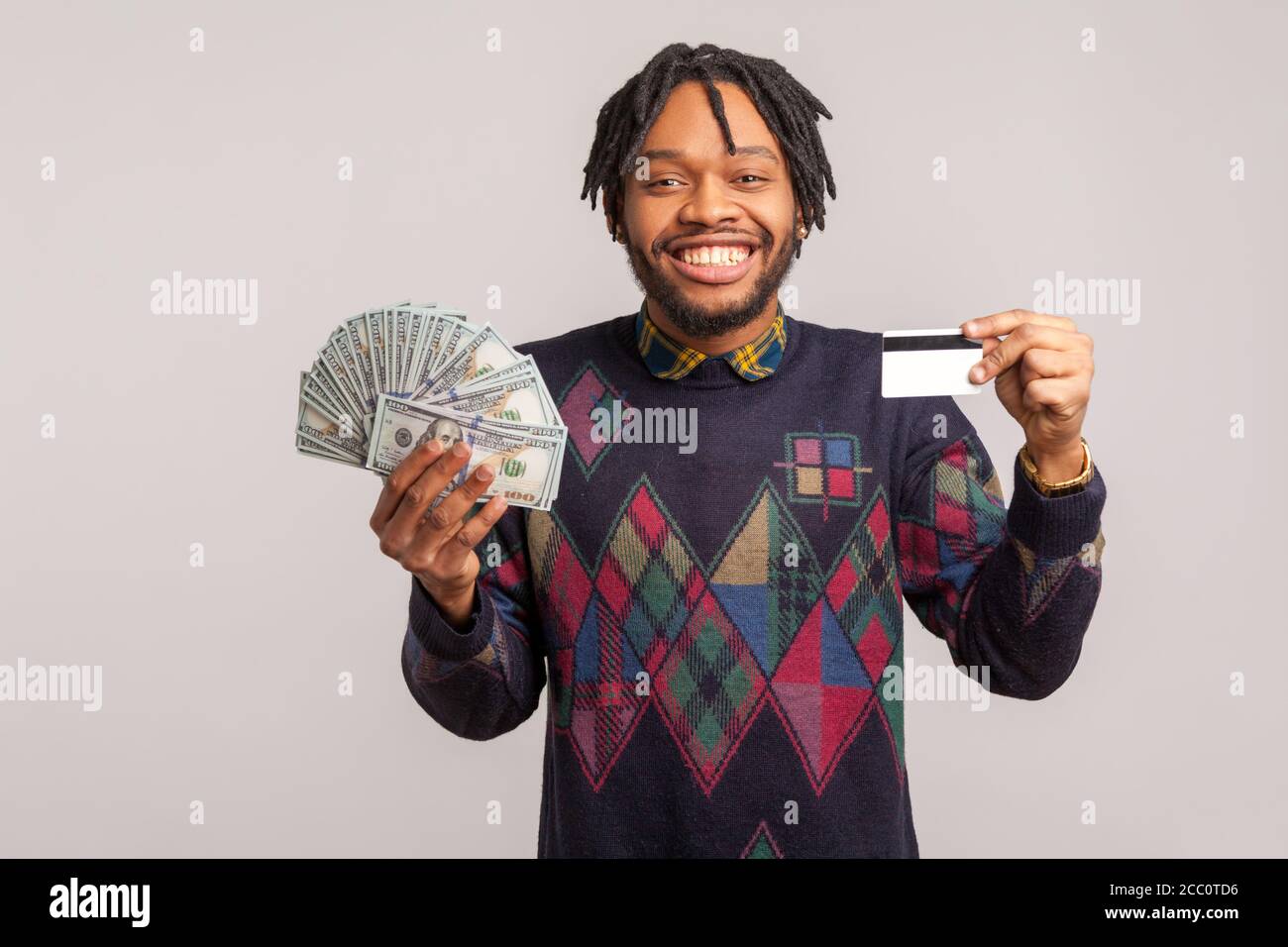 Aufgeregt zufrieden afrikanischen Mann mit Dreadlocks in lässigen Sweatshirt mit Kreditkarte und Charge von Dollar mit toothy Lächeln, leicht Kredit low inte Stockfoto