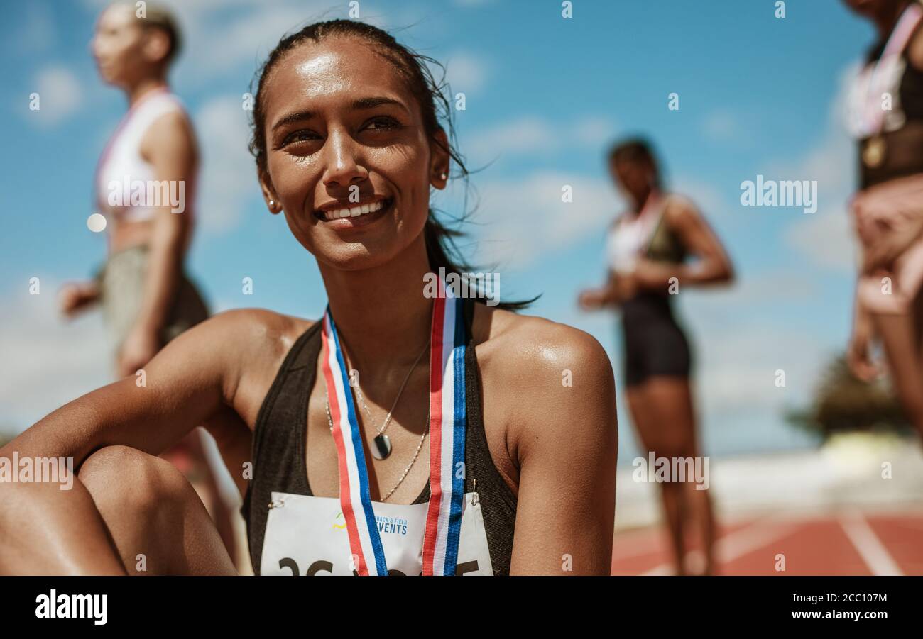 Athletinnen mit Medaille auf der Rennstrecke mit Team im Hintergrund. Sportlerin schaut weg, während sie auf der Leichtathletik-Rennstrecke in sta sitzt Stockfoto