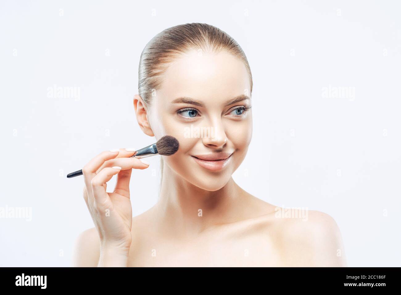 Frauen, Kosmetologie Konzept. Zarte gesunde Europäische Frau gibt Make-up-Unterricht, wendet Kosmetik mit Schönheitsbürste, sieht beiseite mit Lächeln, isoliert auf Stockfoto