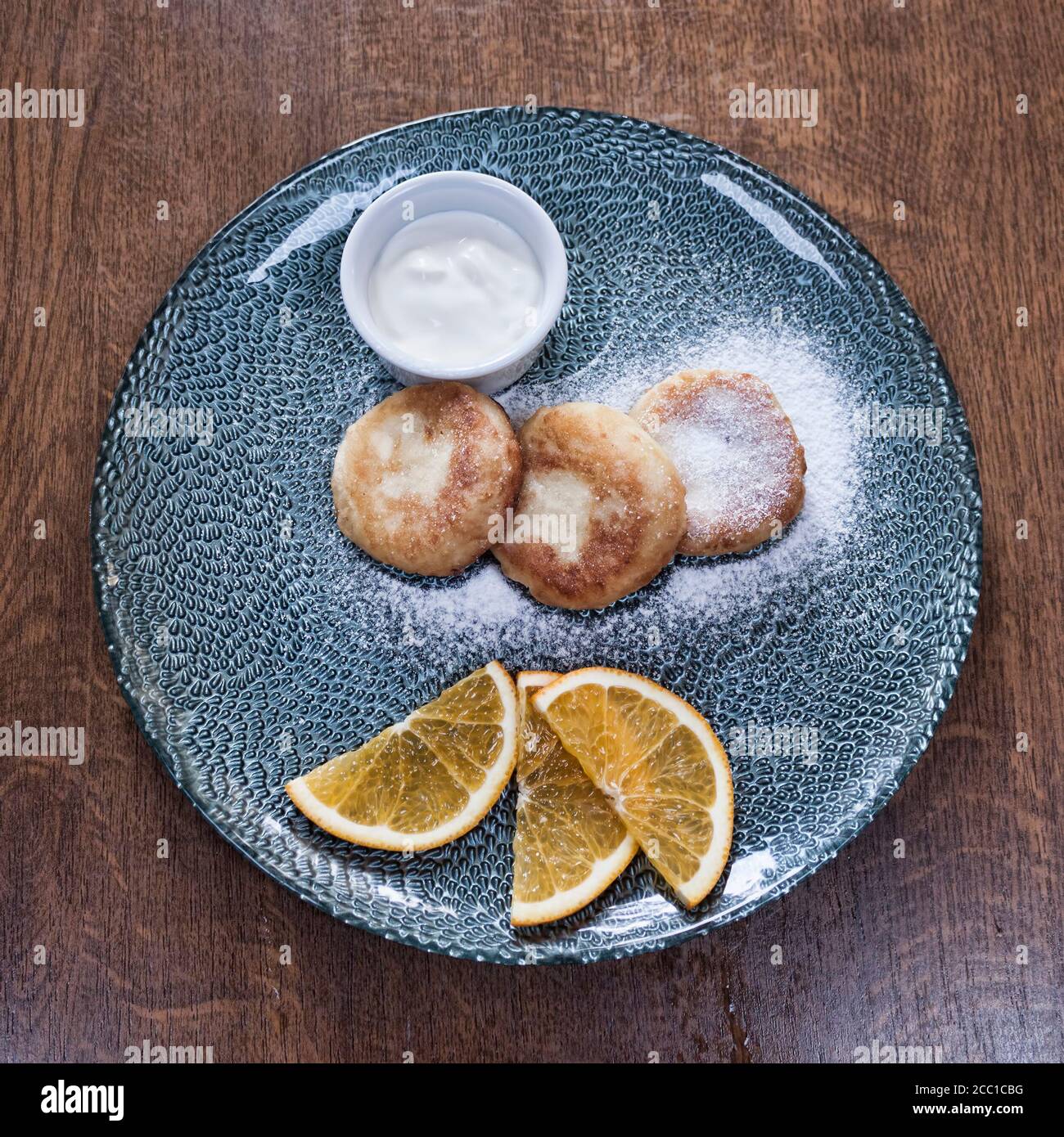 Auf dem Teller sind drei Käsekuchen, bestreut mit Puderzucker, ein Stock mit saurer Sahne und drei Scheiben Orange. Draufsicht Stockfoto