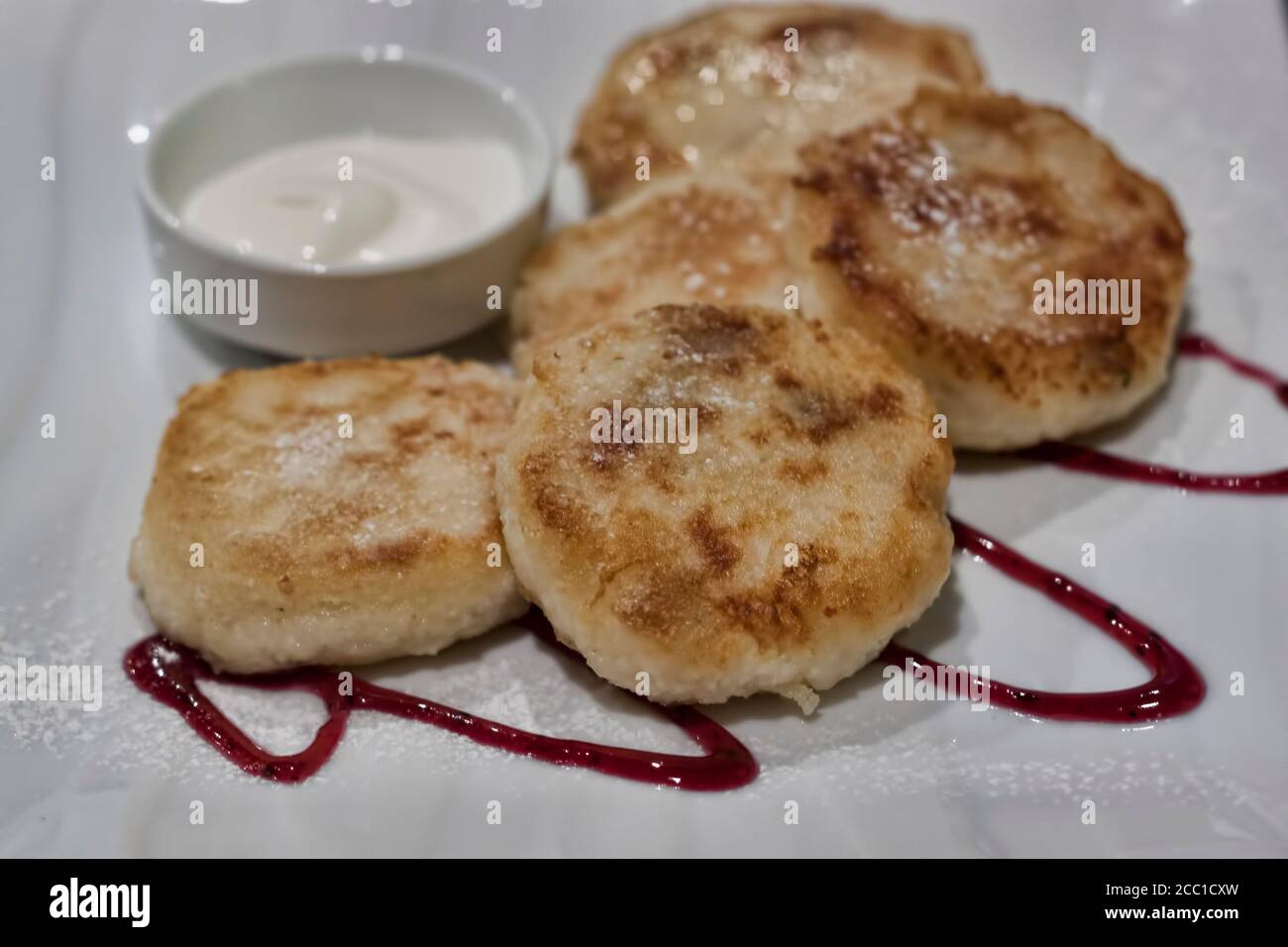Auf dem Teller sind fünf Käsekuchen, bestreut mit Puderzucker, ein Stock mit Sauerrahm und Streifen mit Marmelade bewässert Stockfoto
