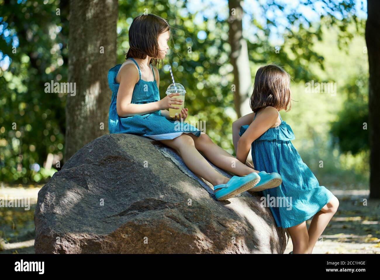 Zwei niedliche kleine Mädchen in Kleidern sitzen auf einem Stein Und Limonade trinken Stockfoto