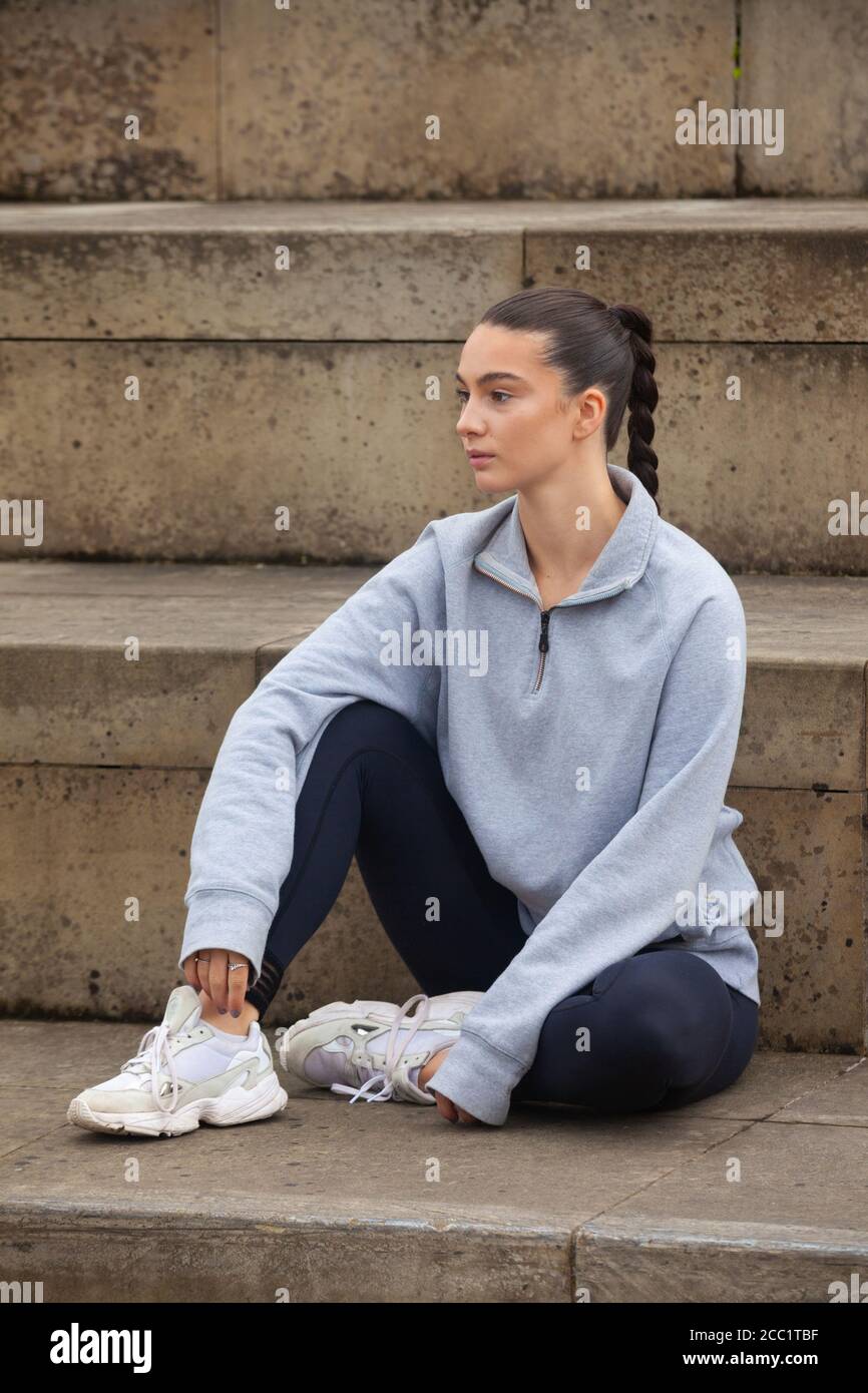 Eine sportliche junge Frau mit Sportkleidung, die vor einer Reihe von Schritten sitzt und bereit für Bewegung ist. Stockfoto