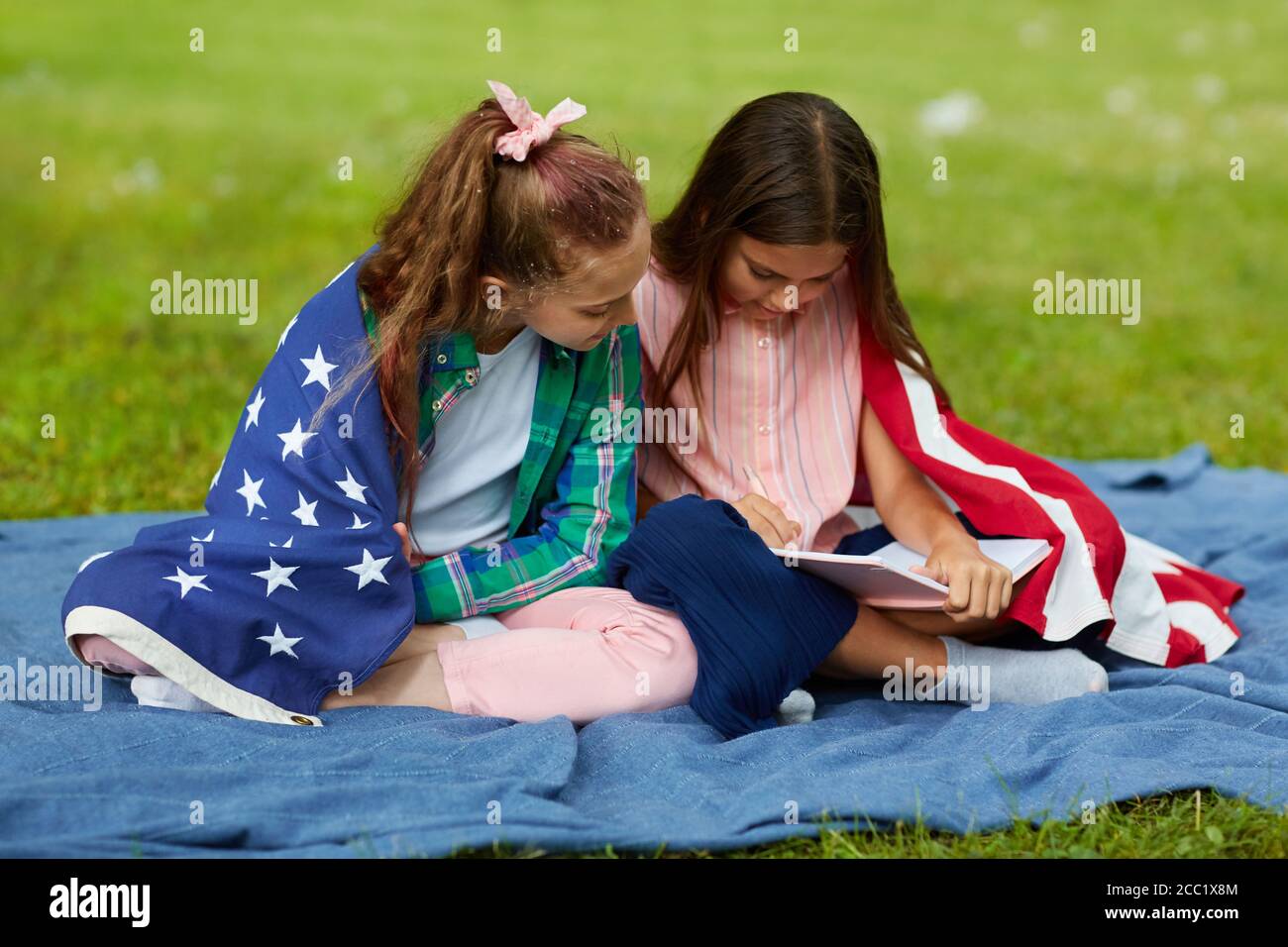 In voller Länge Porträt von zwei niedlichen Mädchen von amerikanischen Flagge bedeckt sitzen auf Picknickdecke im Park und Buch lesen, Platz kopieren Stockfoto