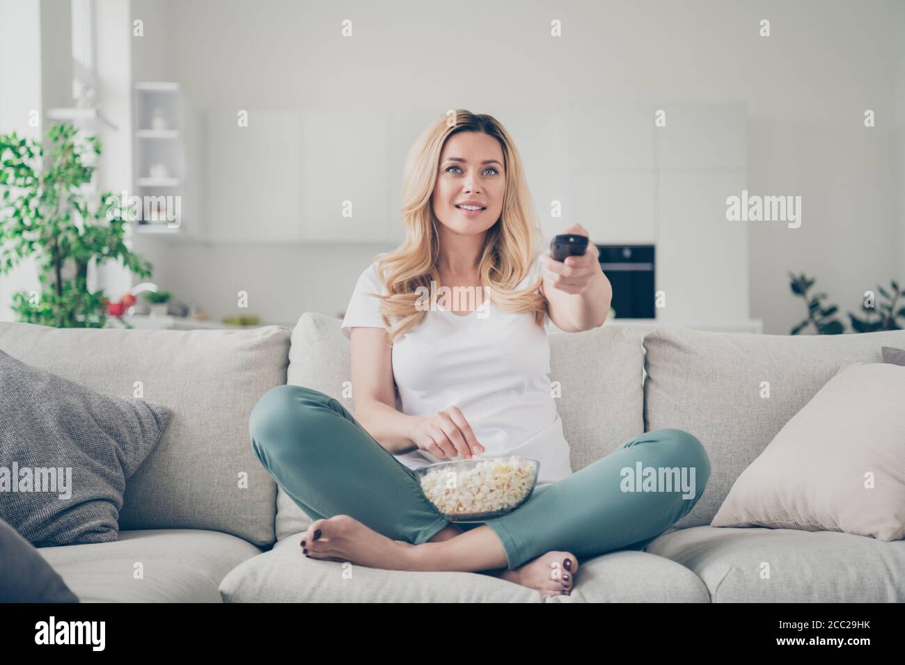 Foto von ziemlich häuslichen schönen Dame sitzen Couch essen Popcorn Fernbedienung ändern Sender Fernsehen Wochenende Quarantäne zu Hause bleiben Gute Laune Beine Stockfoto